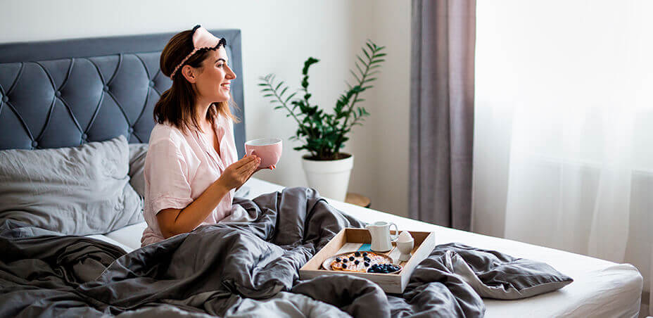 Brunette lady having coffee  sitting in a bed with grey and white bedding and grey upholstered headboard